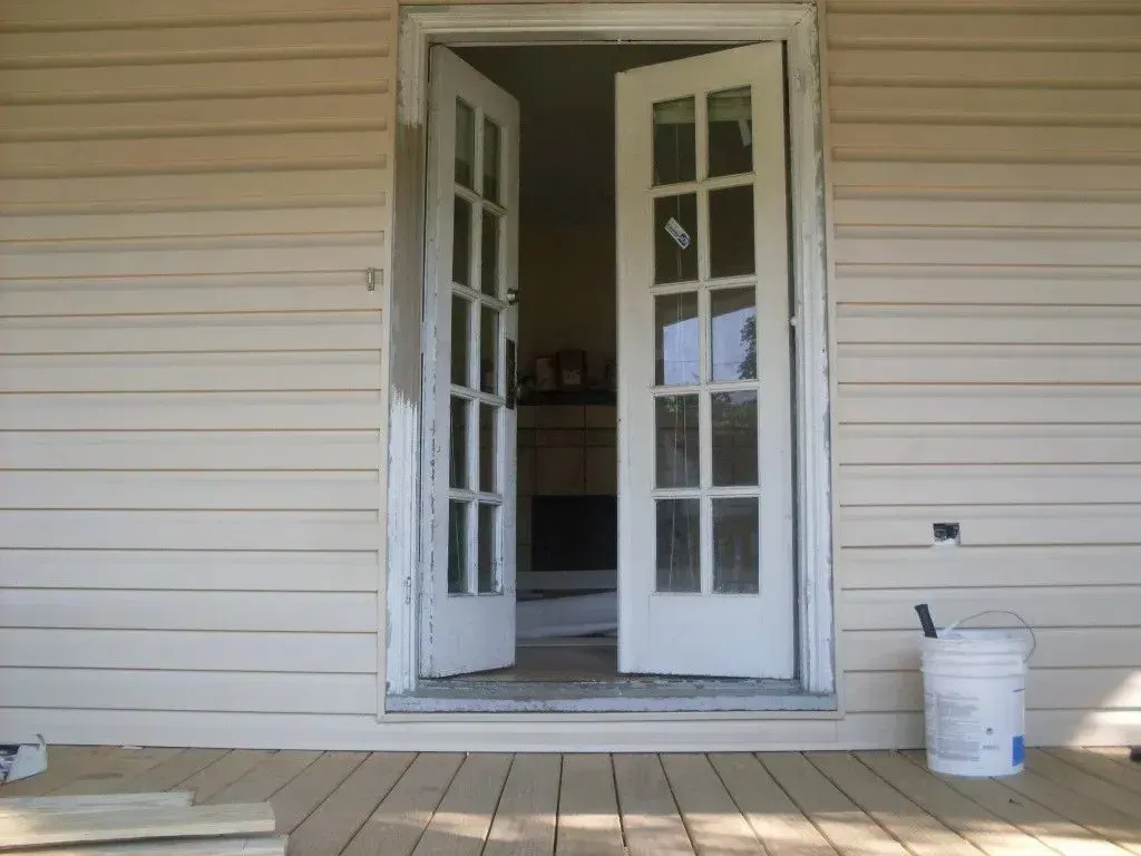 A bucket of paint sits on a porch next to an open door