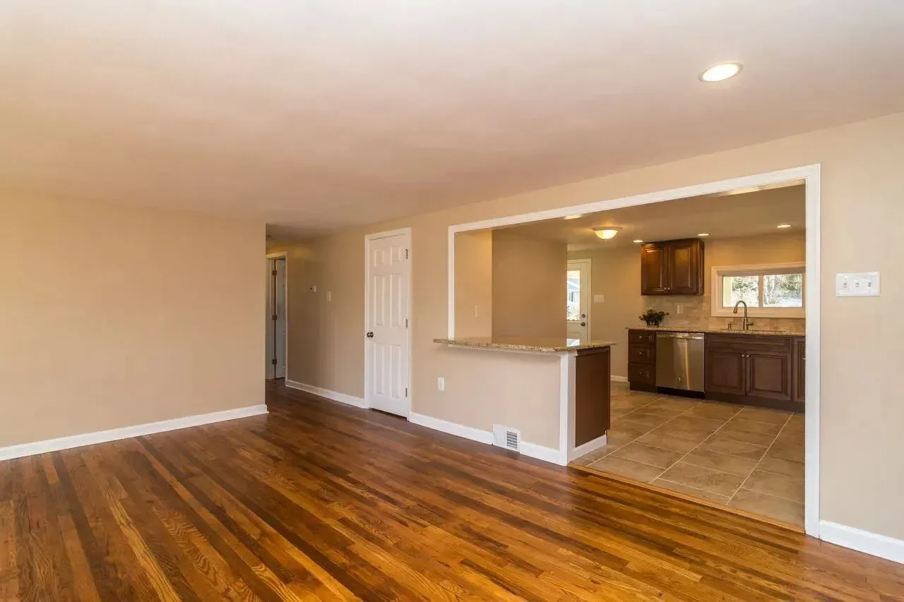 An empty living room with hardwood floors and a kitchen in the background.