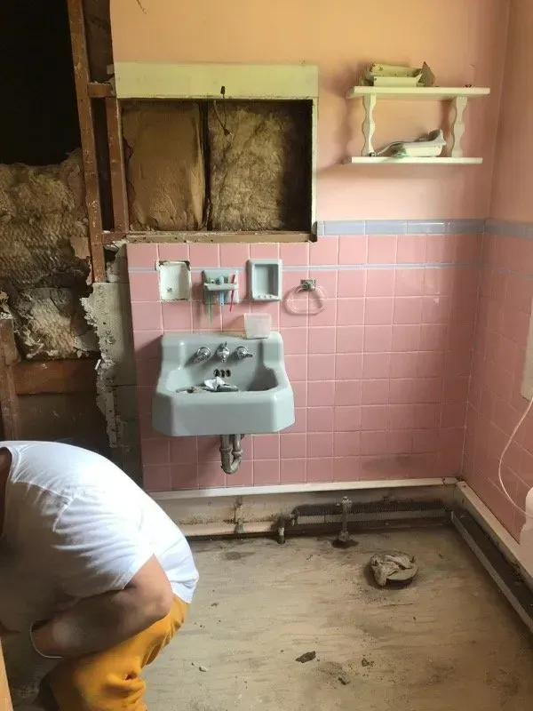 A man is squatting in a bathroom with pink tiles and a sink.