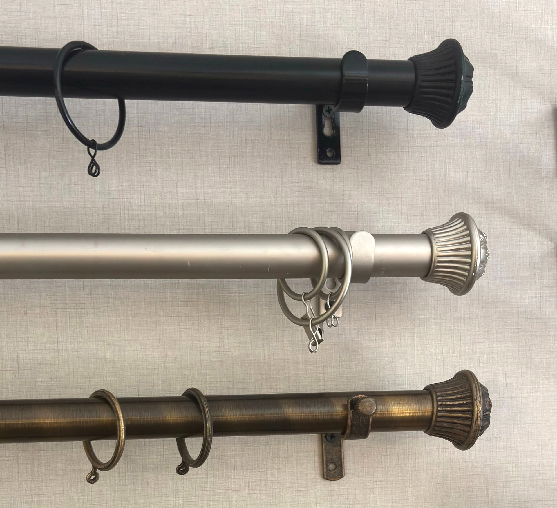Tres barras de cortina: negra, plateada y bronce. Cada una tiene remates y anillas decorativas.