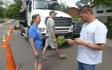 Three people stand on a street near a large white dump truck and traffic cones, with one person looking at a tablet.
