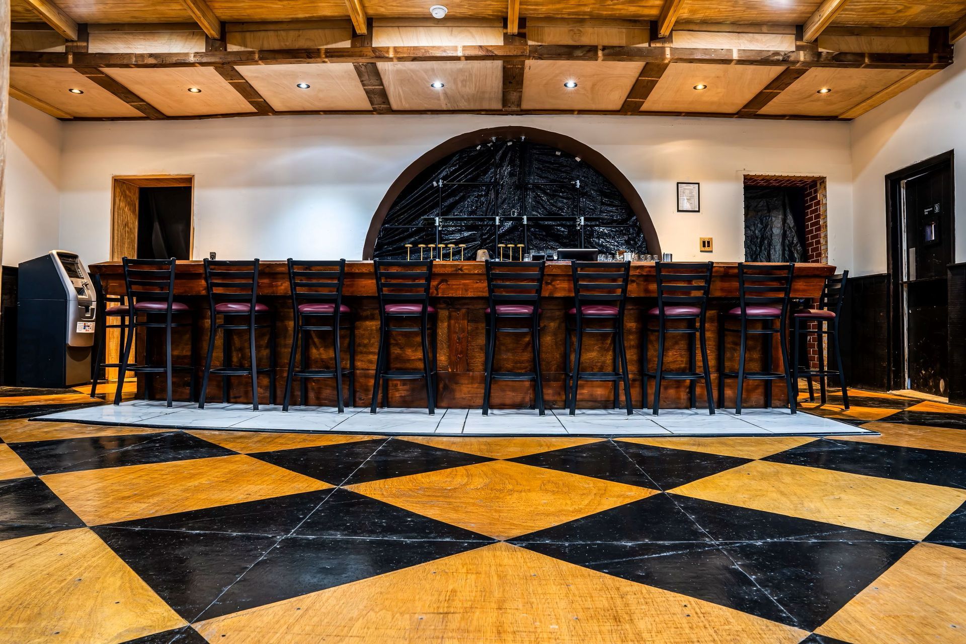 A long wooden bar with stools in a room with a checkered floor.