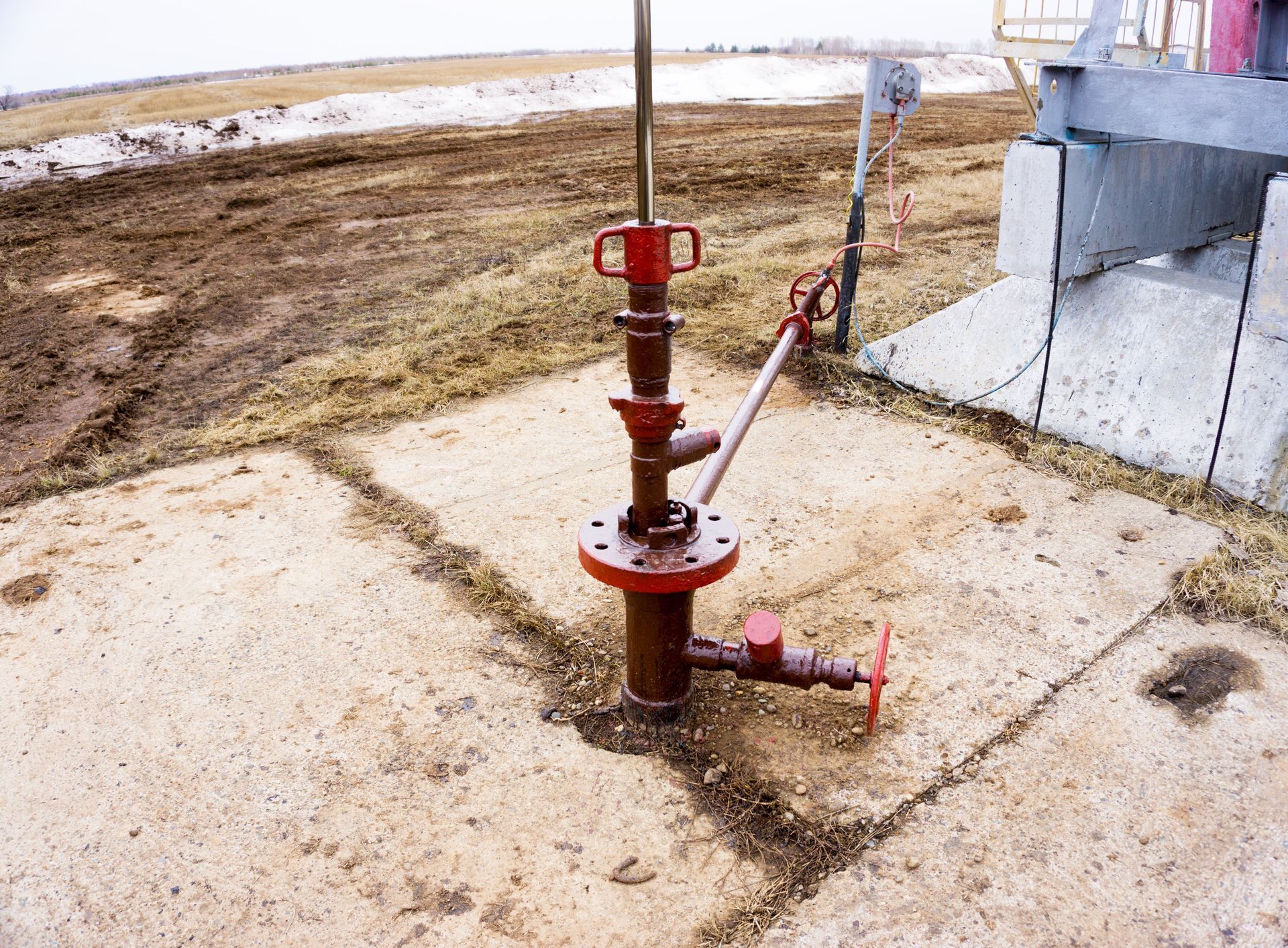 Red wellhead equipment on concrete pad, likely for oil or gas extraction in a muddy field.
