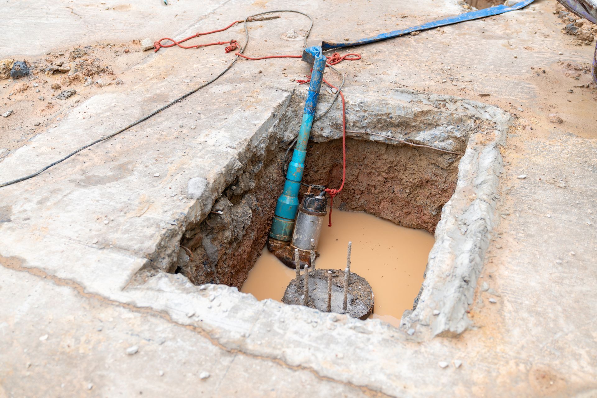 Concrete pit with pump submerged in murky water; blue pipe, red and black cords.