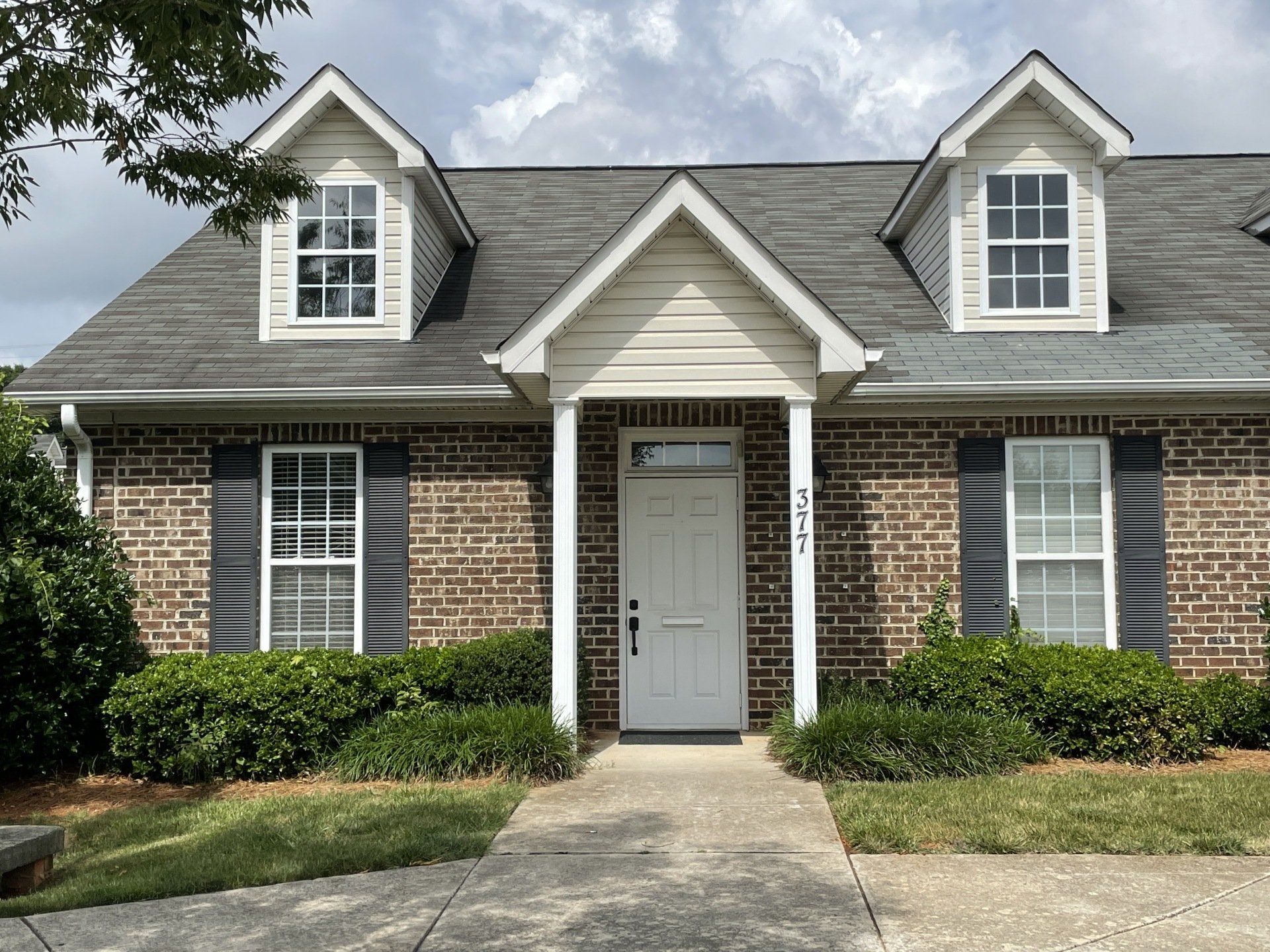 Brick townhouse with white door, three dormer windows, and black shutters, set in a yard.