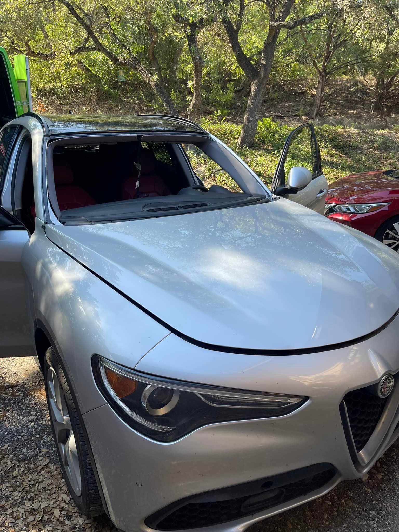 A silver car with a broken windshield is parked in the woods.