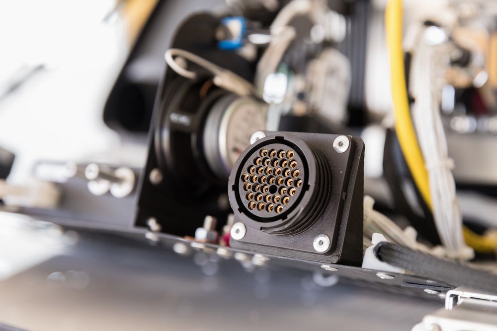 Close-up of a circular electrical connector on a black metal device with wires in the background