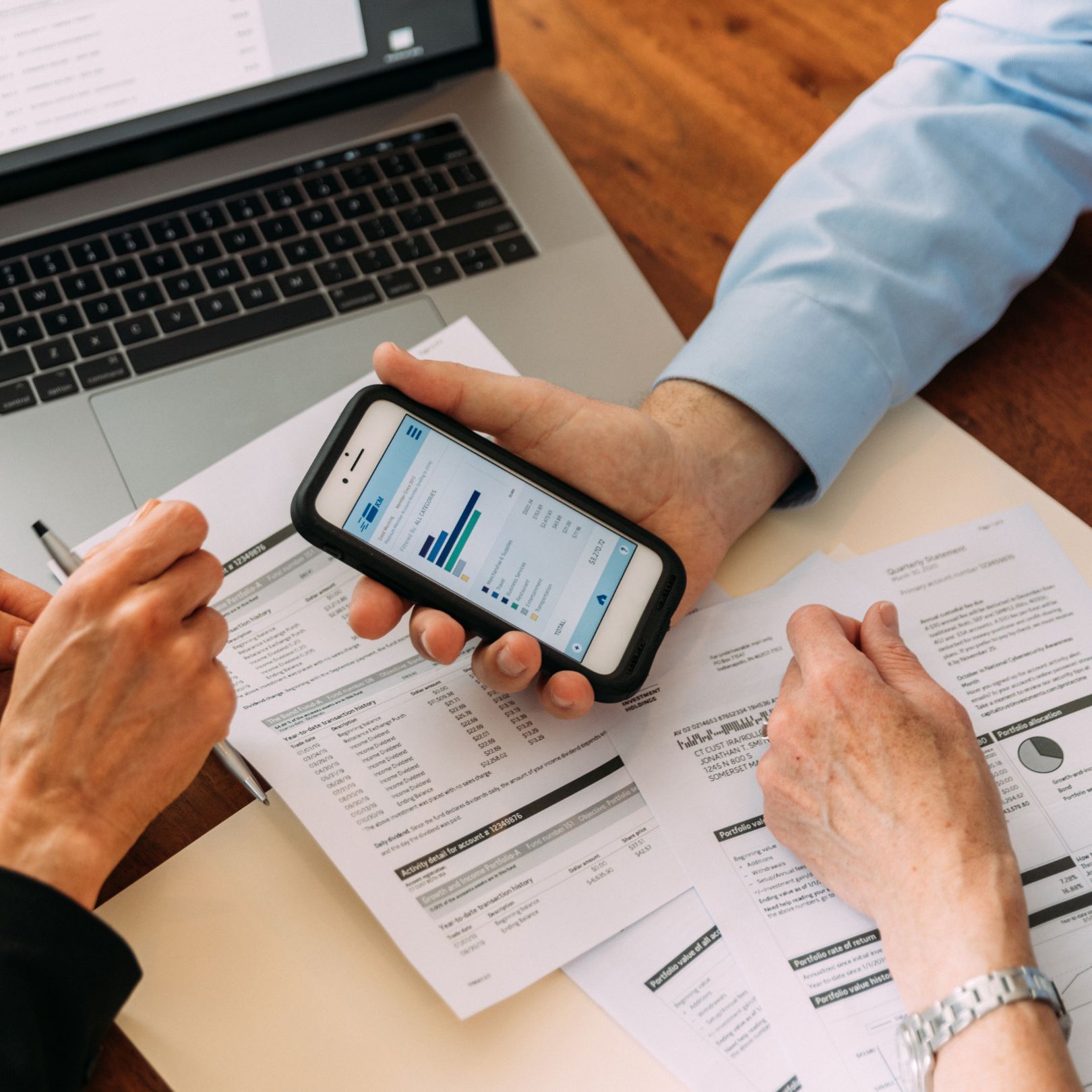 Hands examining financial documents, a laptop, and a smartphone displaying a graph.