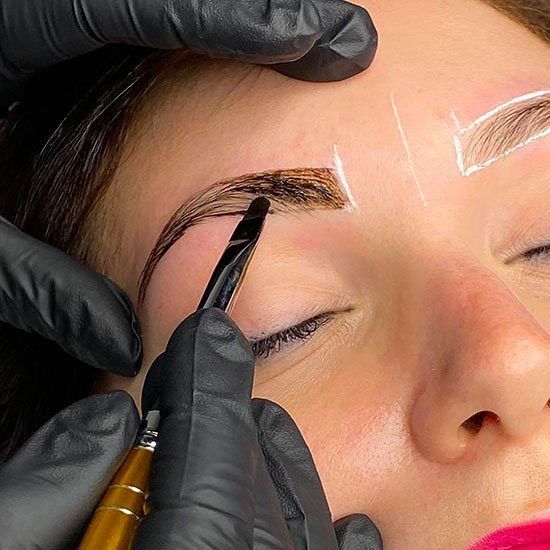 Procedura di microblading delle sopracciglia: occhio chiuso, mani guantate e uno strumento che applica il pigmento all'interno di uno stencil.