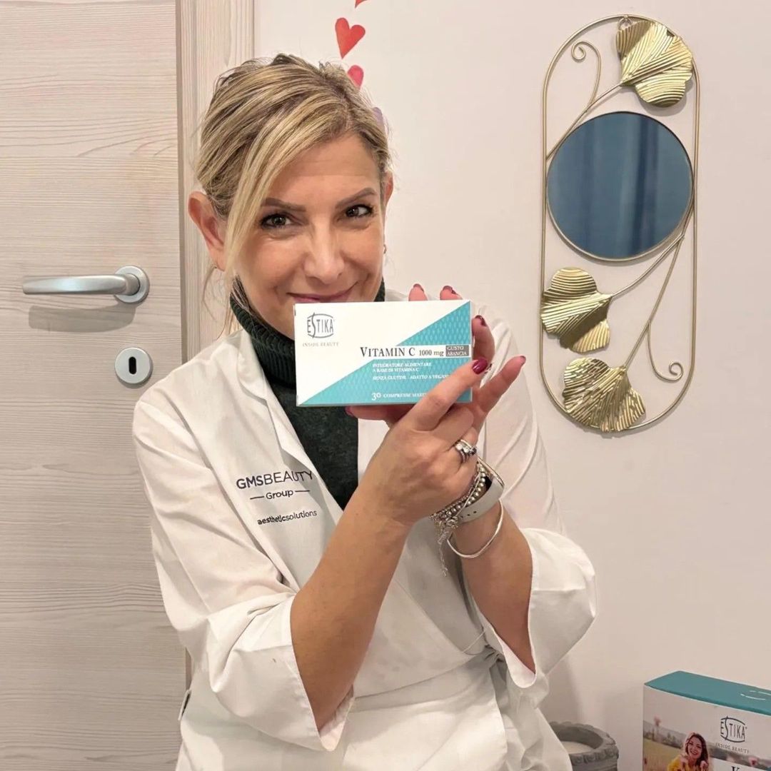 Donna in camice da laboratorio che tiene in mano una scatola di prodotti, sorridente. Ambientazione interna con decorazione murale.