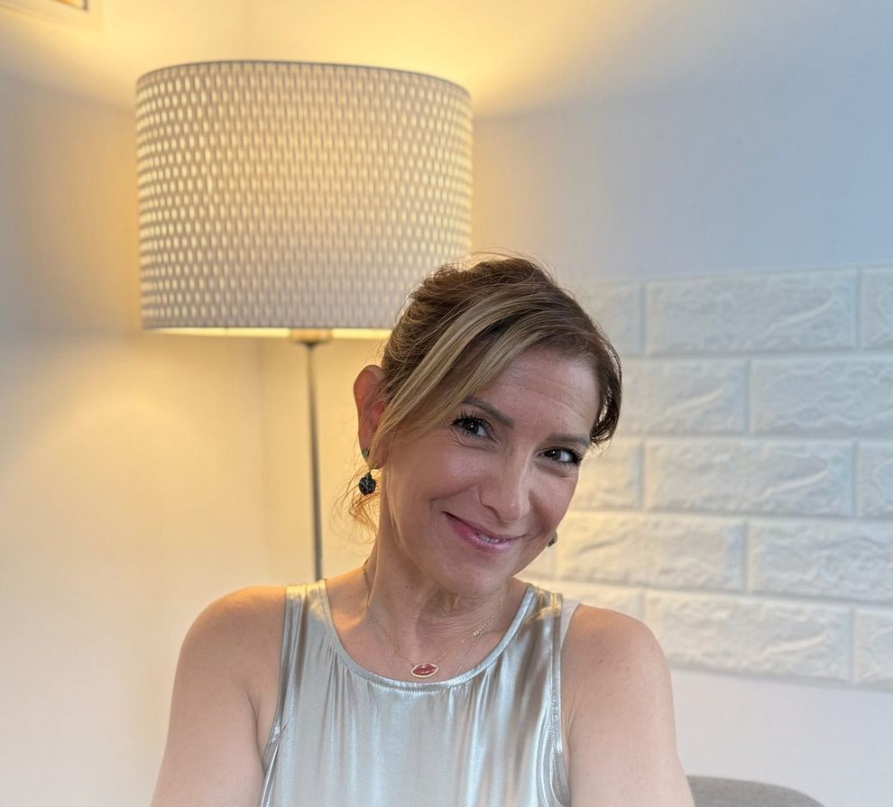 Una donna sorride, seduta all'interno, con indosso un top argentato, accanto a una lampada e a un muro di mattoni bianchi.