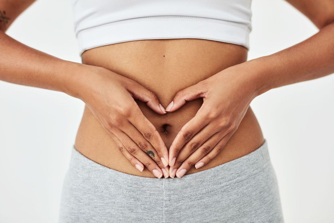 Donna che si tiene le mani a forma di cuore sopra l'addome, indossa un top bianco e pantaloni grigi.