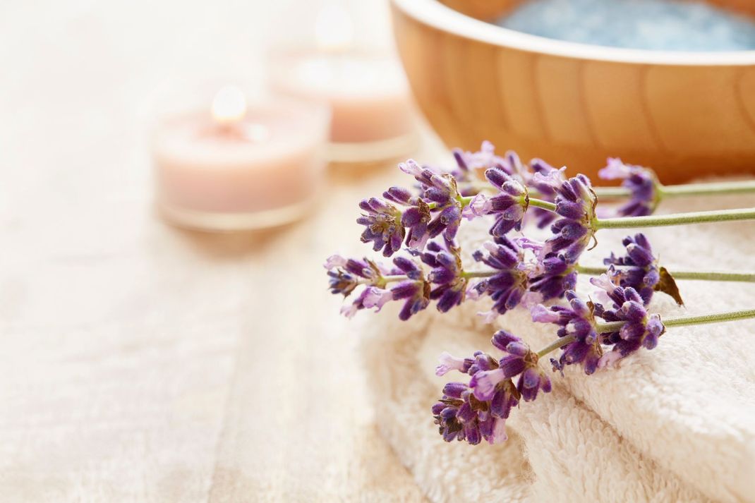 Rametti di lavanda su un asciugamano bianco, con candele e una ciotola di legno con contenuto blu.