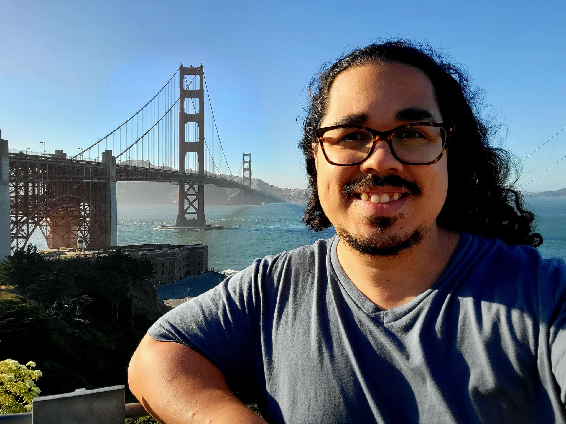 A man with long wavy dark hair in a blue tshirt and glasses standing in front of the Golden Gate Bridge wearing glasses smiling at the camera