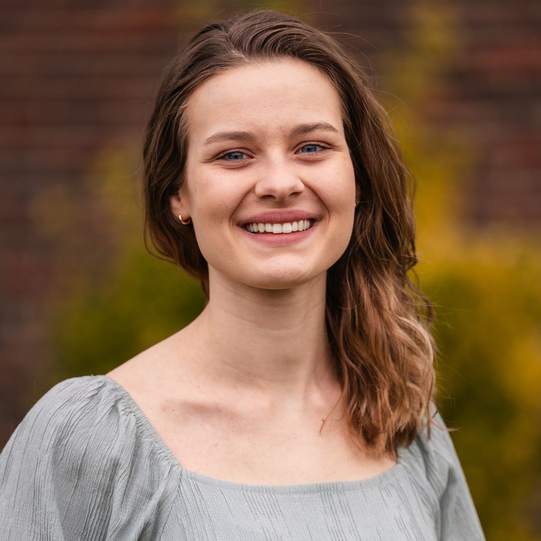 Woman with long brown hair, smiling at the camera, wearing a light blue top. Background includes brick and greenery.