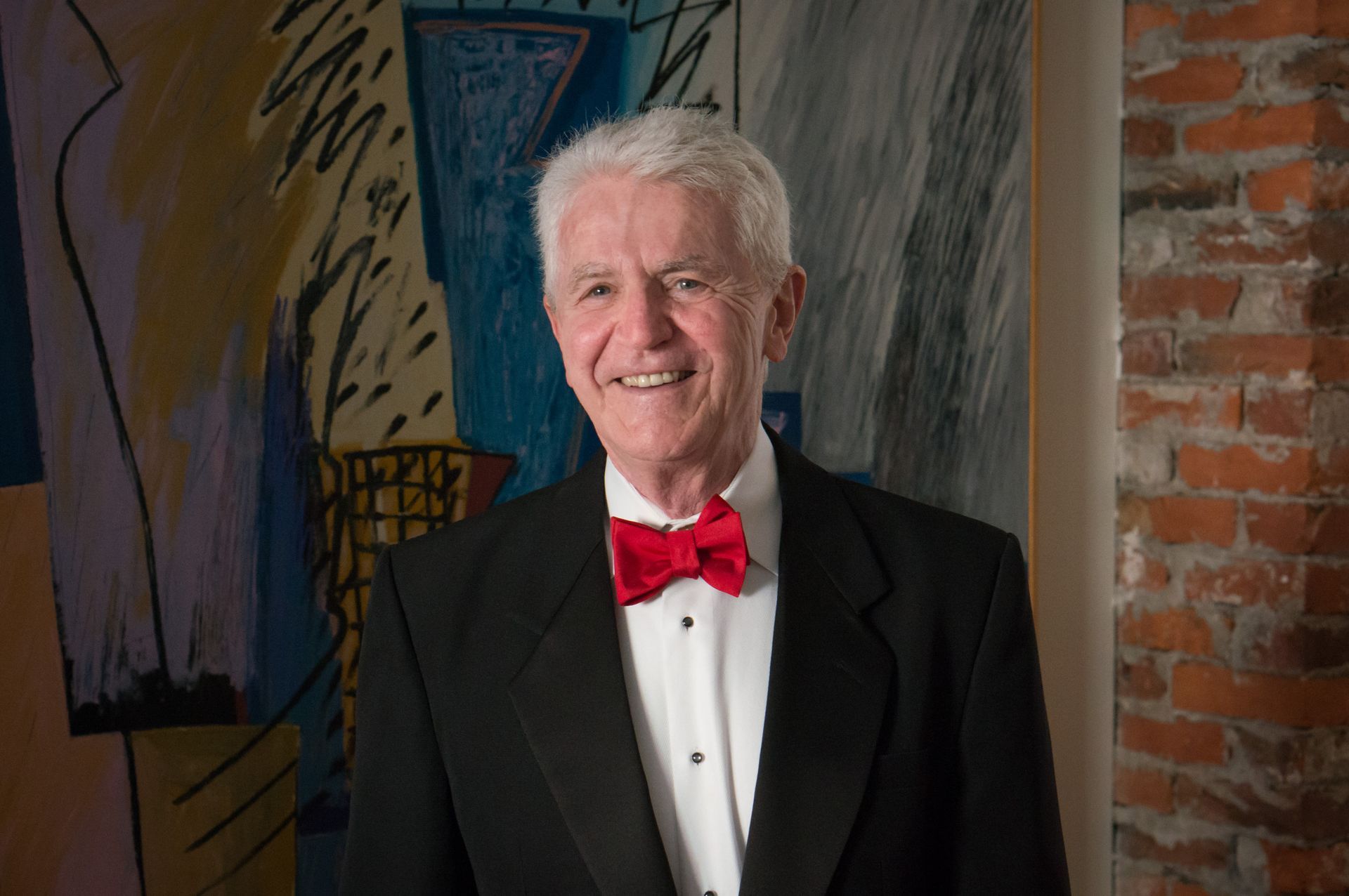 Man in tuxedo with red bow tie smiles in front of a painting and brick wall.