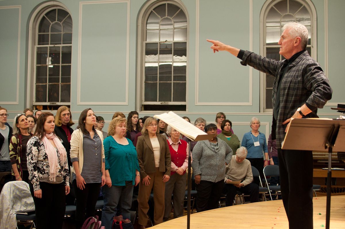 Choir conductor directing a group of singers in a light-blue-walled room. The conductor points, choir sings.