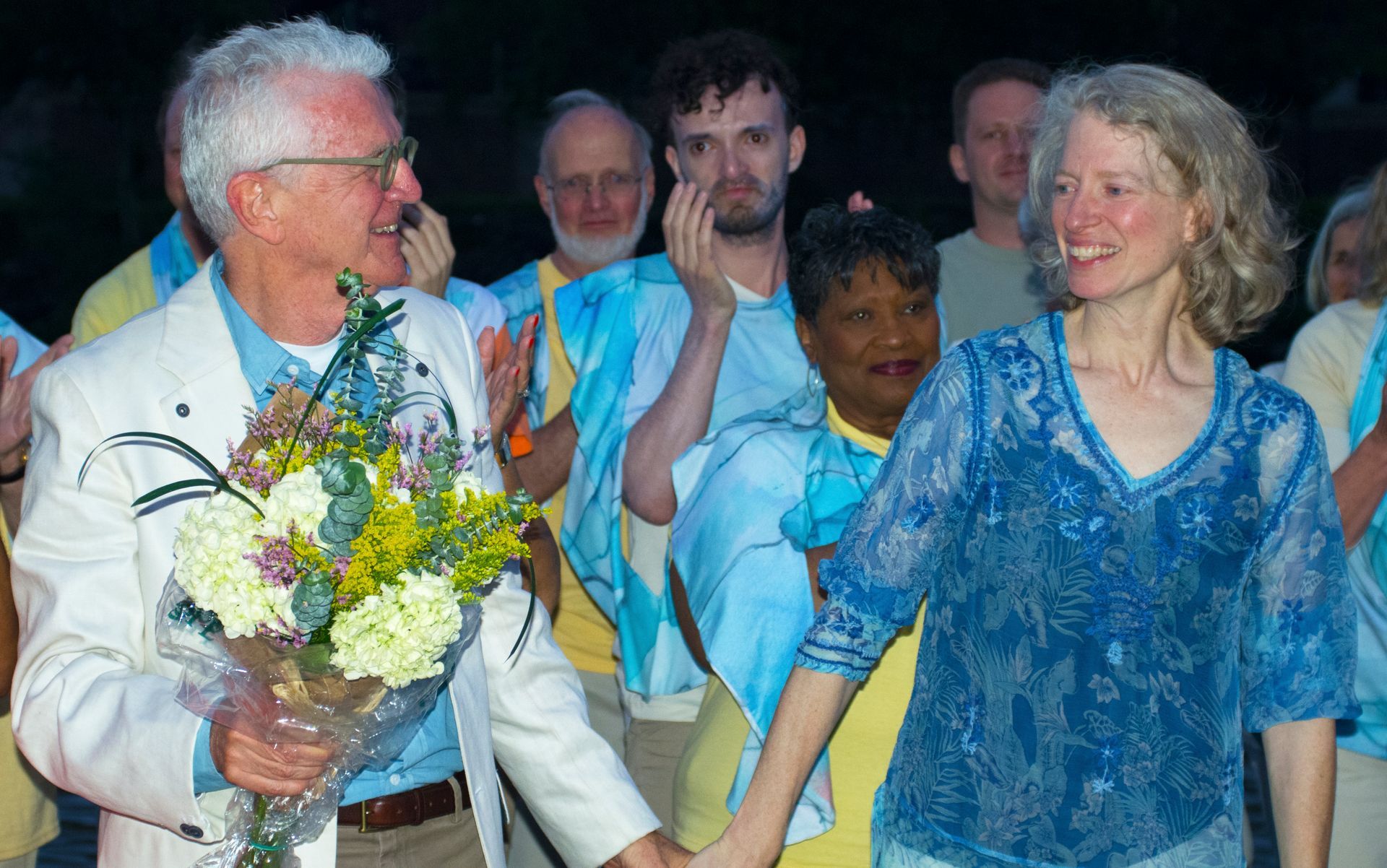 Man gives flowers to woman, both smiling, surrounded by people in blue, outdoor setting.