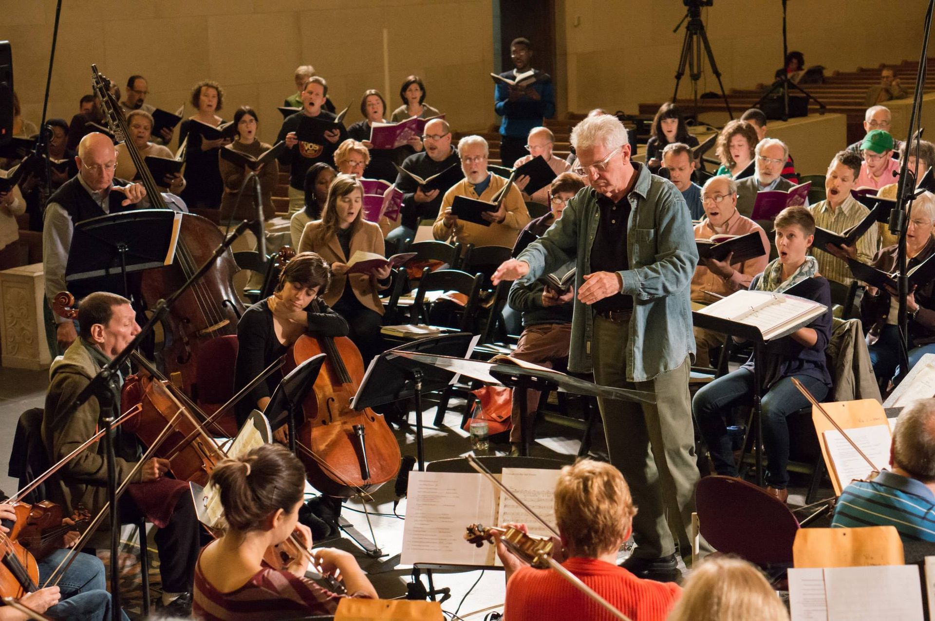 An orchestra conductor leads a performance, gesturing with a baton. Musicians play instruments with a choir in the background, performing in a large hall.