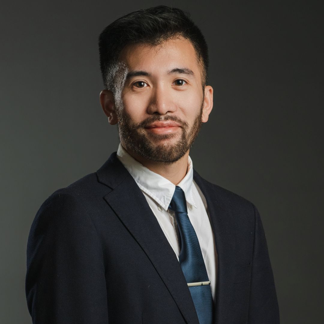 Asian man in a suit and tie smiles at the camera against a dark gray background.