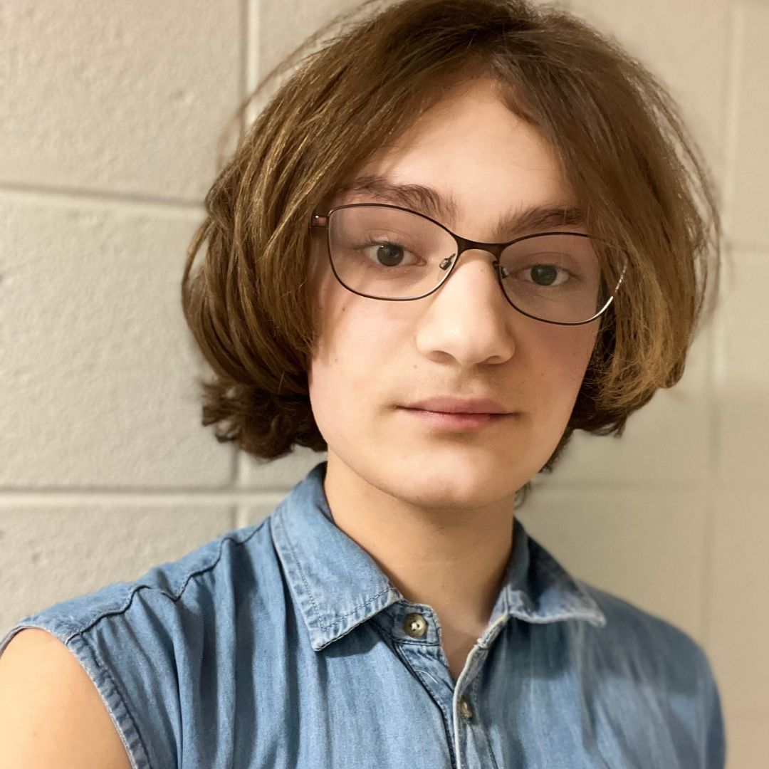 A person with light brown hair and glasses wearing a sleeveless denim shirt, standing in front of a white brick wall.