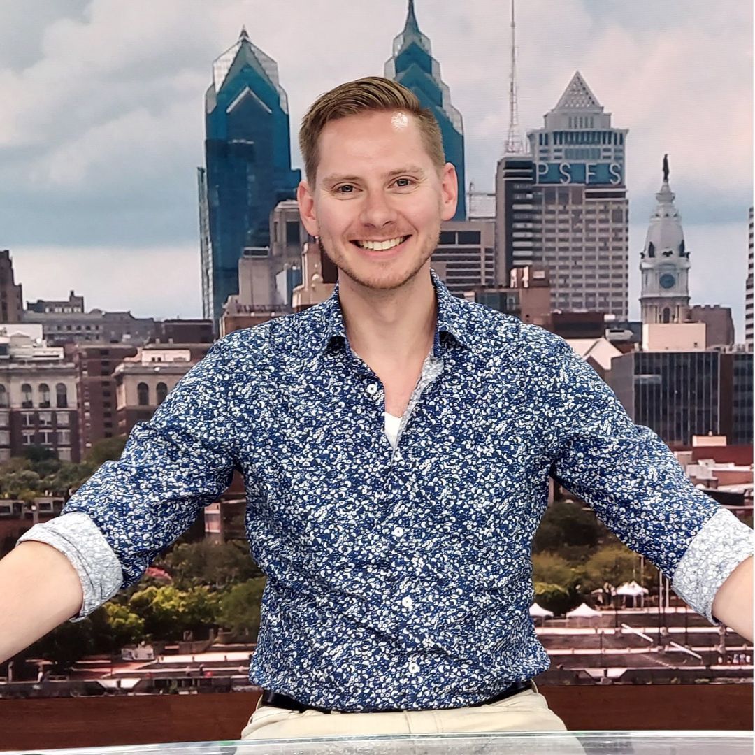 Man with fair skin in a blue floral shirt smiles, arms outstretched, in front of a Philadelphia skyline.