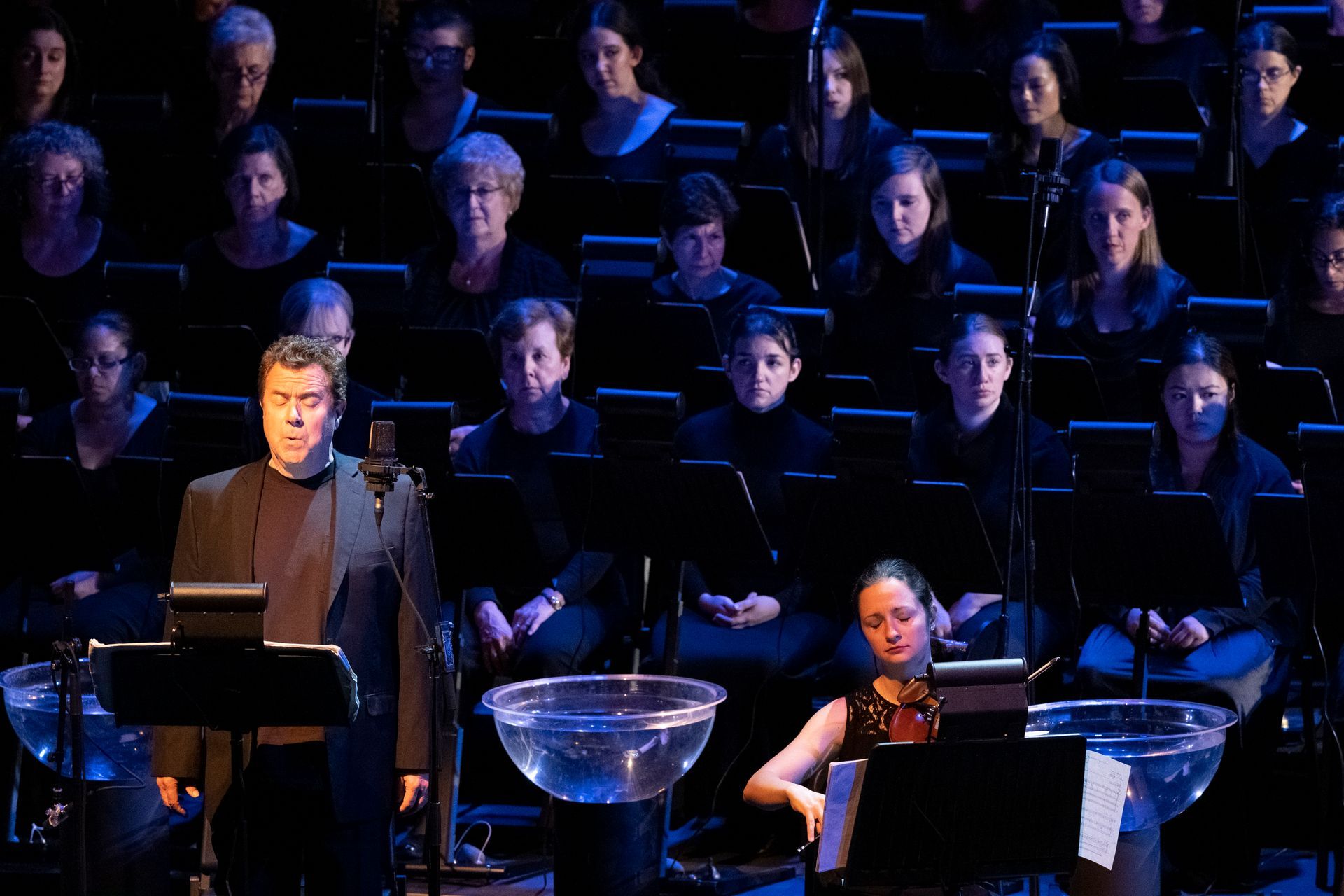 A man is standing in front of a large orchestra while a woman sits at a piano.