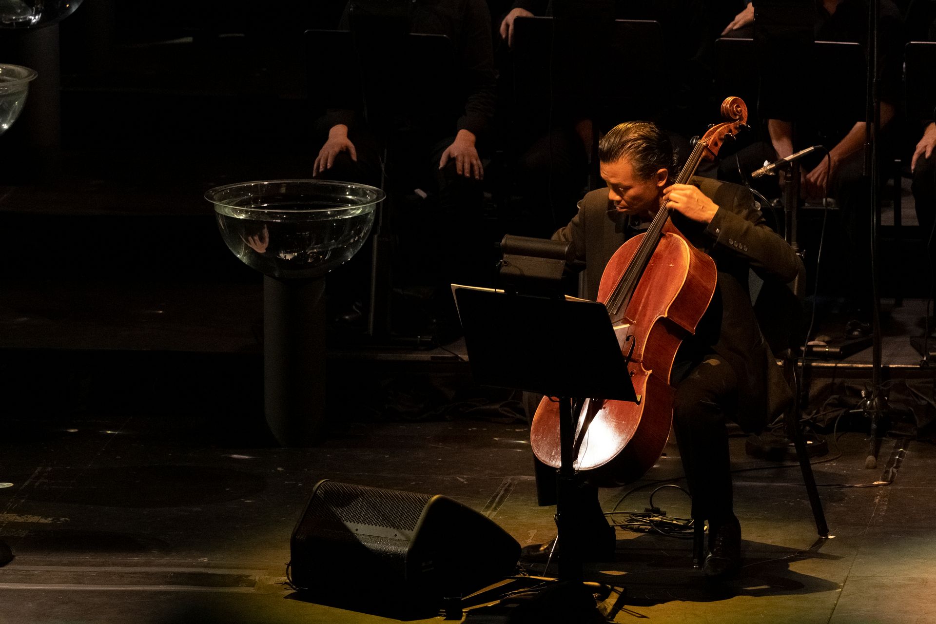 A man is playing a cello on a stage in front of an orchestra.