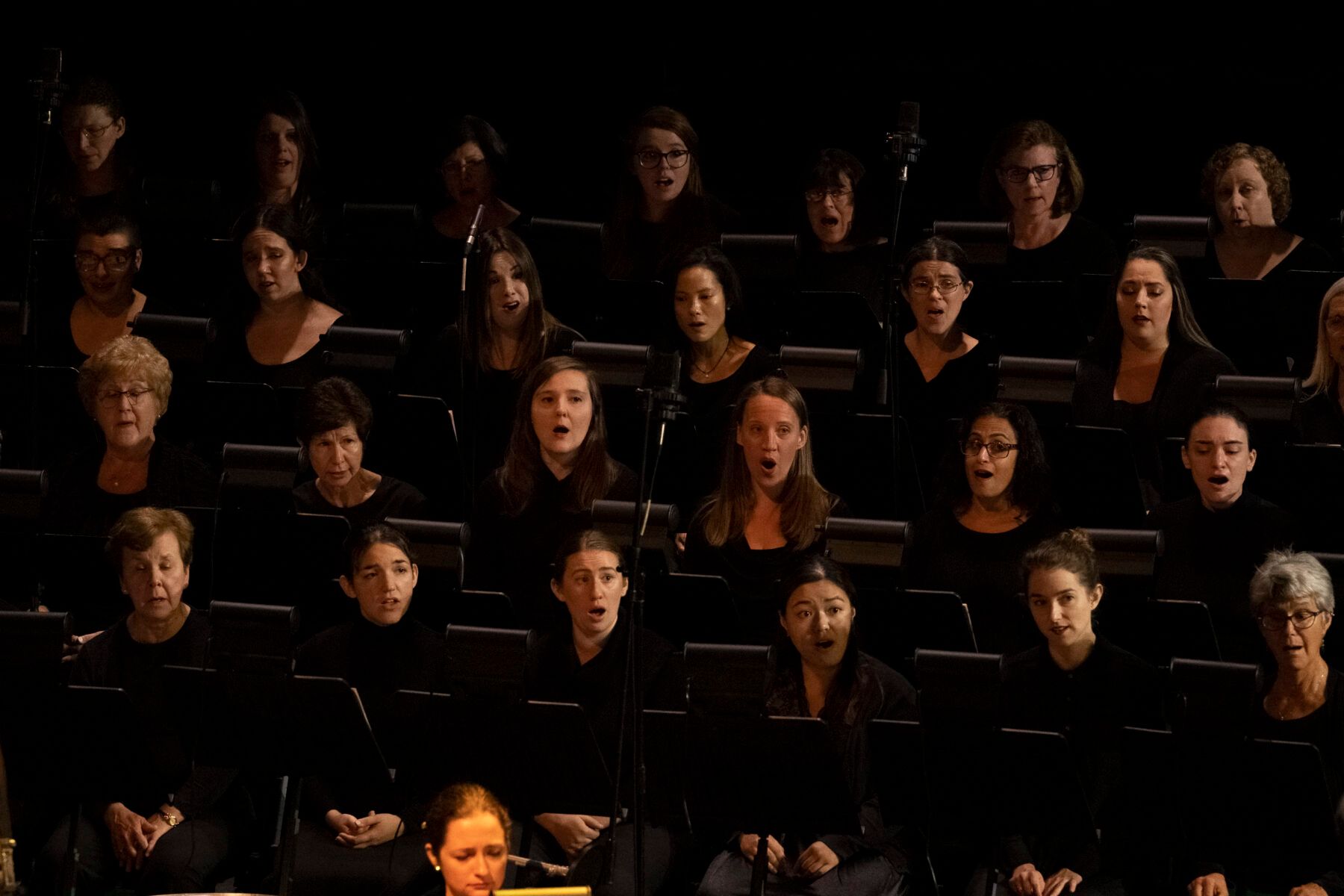 A large group of people are singing in a choir in a dark room.