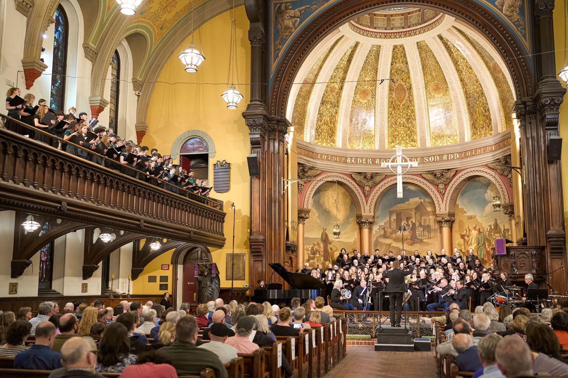 Choir performing in a church. A large crowd watches from pews. The church has a dome ceiling and ornate decorations.