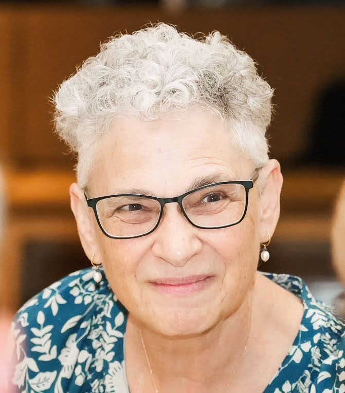 Woman with short, gray hair and glasses, wearing an orange top, smiles at the camera. She stands in front of a gray wall, a lanyard around her neck.