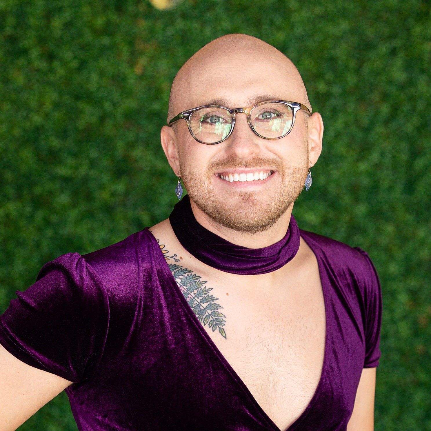 A bald man wearing glasses and a purple dress is smiling for the camera.