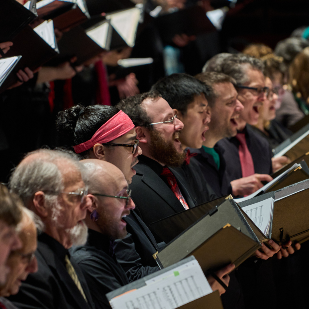 Choir members singing from sheet music.  Men and women are shown, some with open mouths, in a concert hall setting.