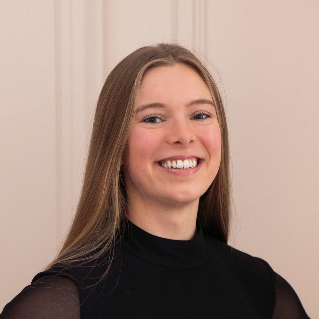 Smiling young woman with long brown hair, wearing a black top against a white background.