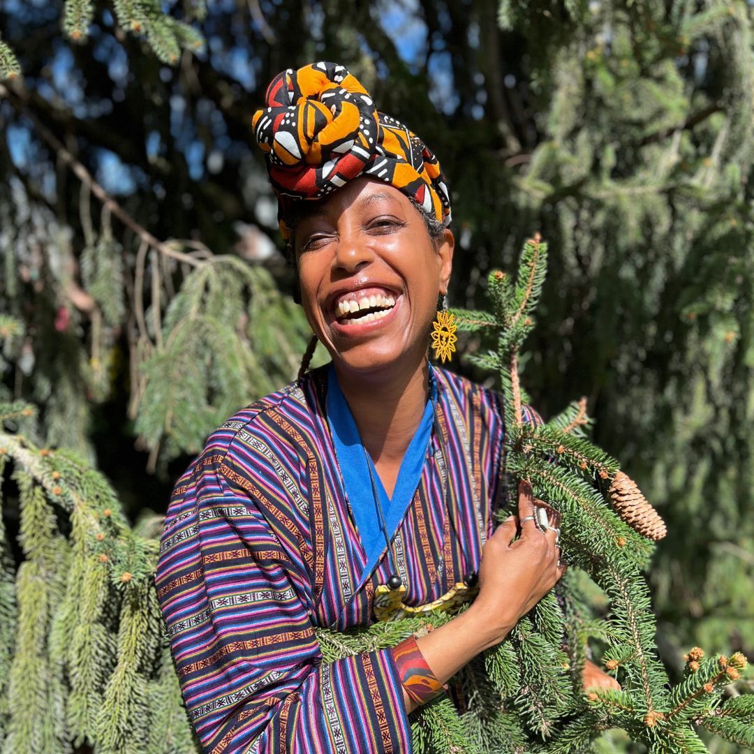 Woman with headwrap smiling, holding a pine branch, against a backdrop of a lush evergreen tree.