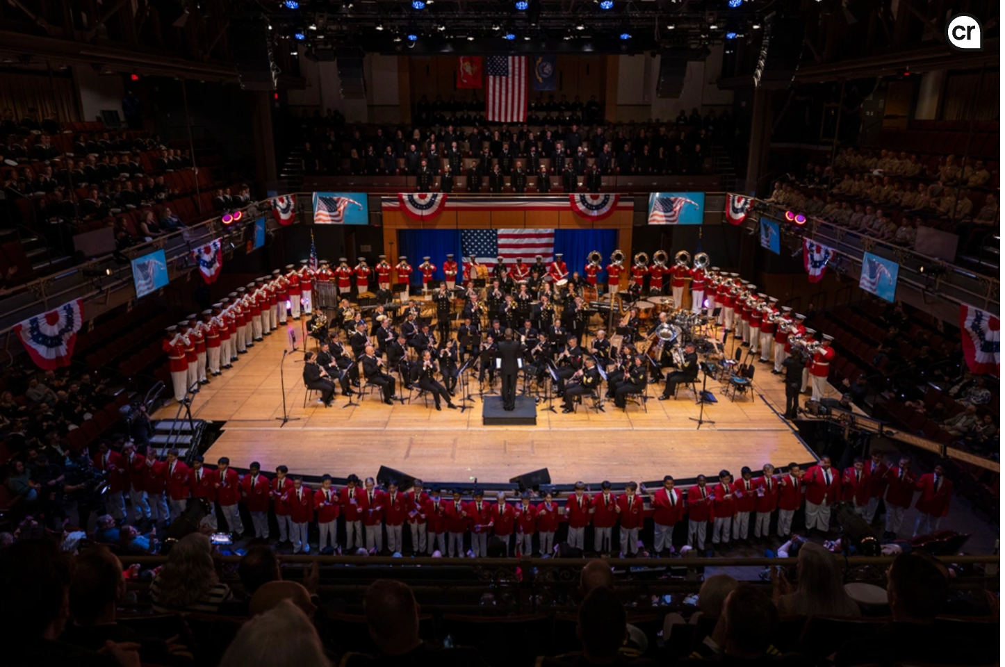 Audience watches a band and choir perform on stage, American flag in the background, red and white colors.