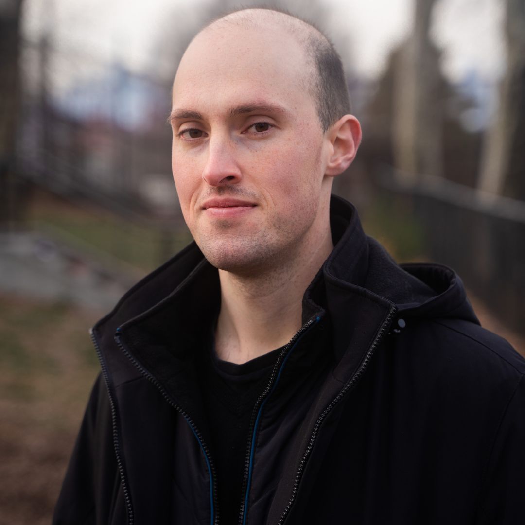 Bald man in a black jacket, smiling, outdoors. Blurred background of trees and fence.