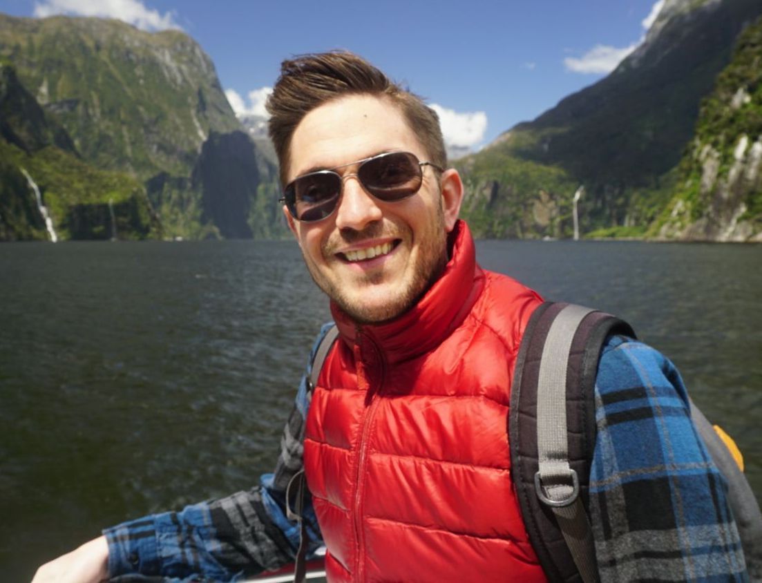 A man wearing sunglasses and a red vest is standing in front of a lake.