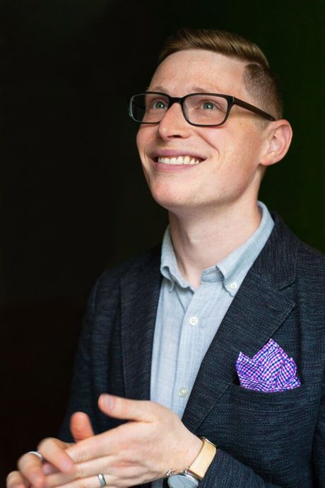 A man wearing glasses and a purple pocket square is smiling.