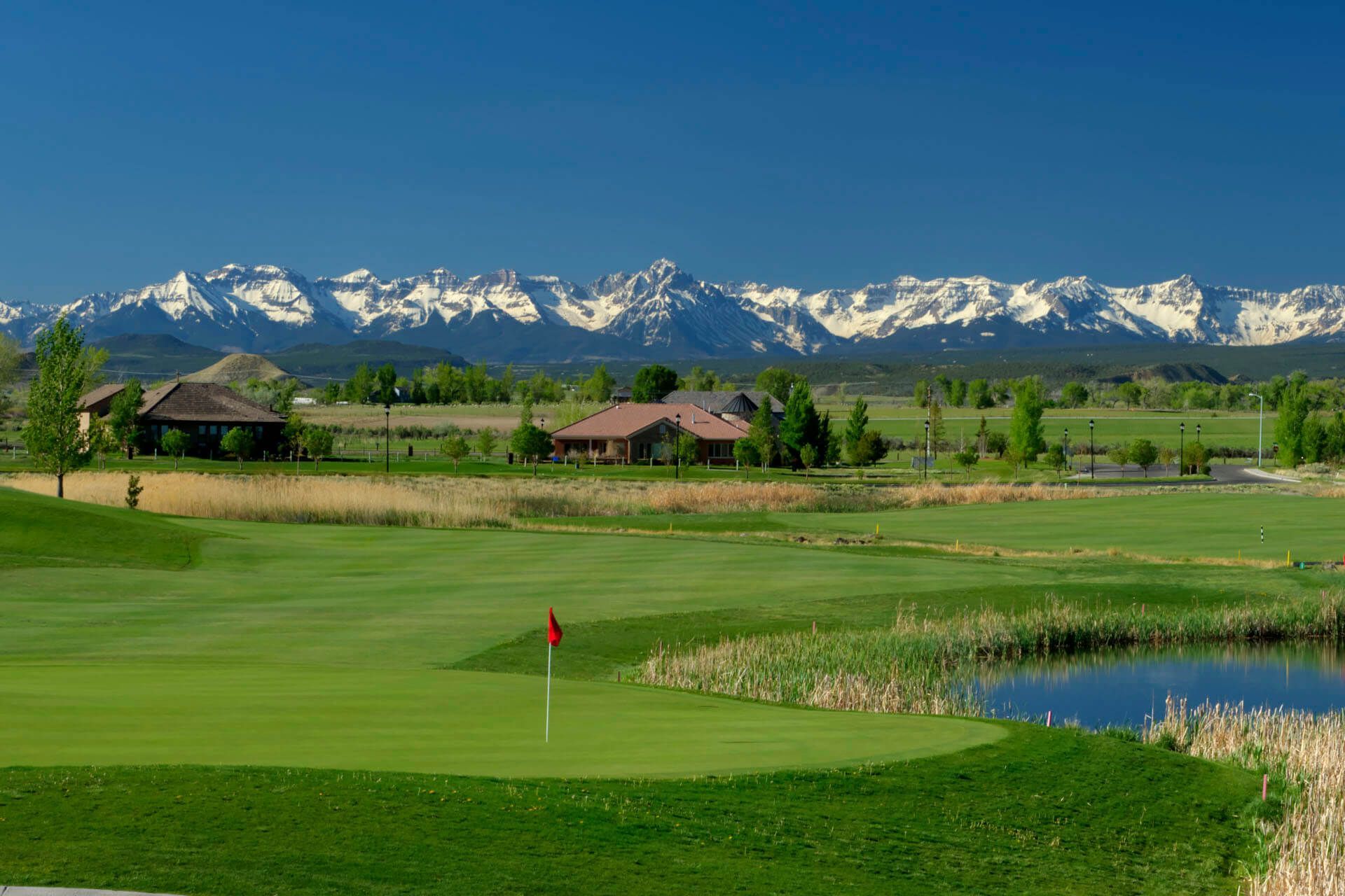 Green golf course with red flag, pond, houses, and snow-capped mountains under a blue sky.