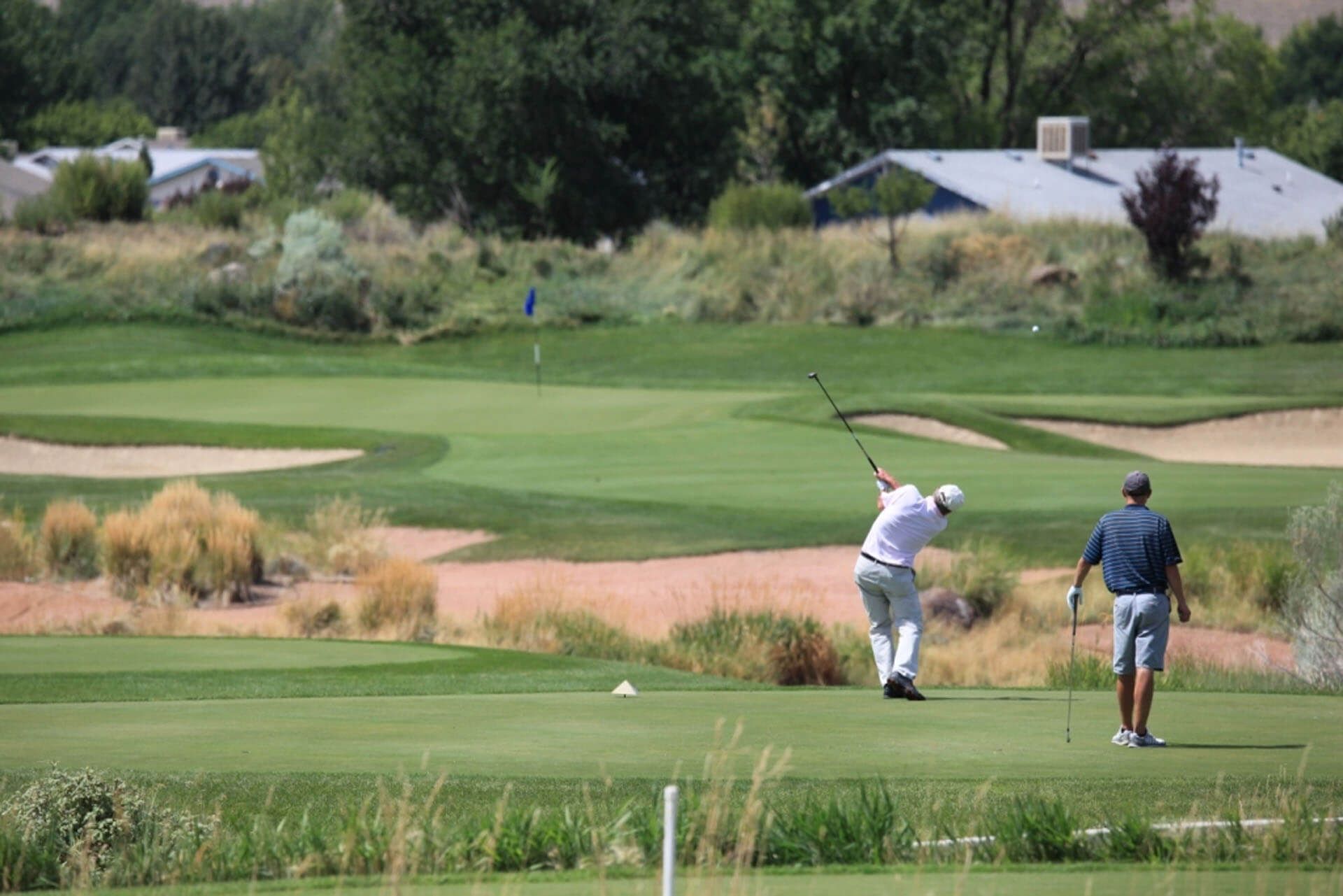 Golfer swinging club on green golf course; another watches. Light green grass, sand traps, and blue sky.