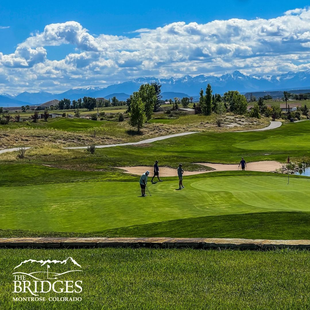 Golfers on a green at The Bridges in Montrose, Colorado, with mountains and cloudy blue sky in the background.