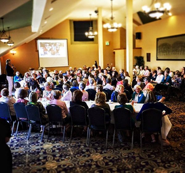 Large group of people seated at tables in a banquet hall, watching a presentation on a screen.