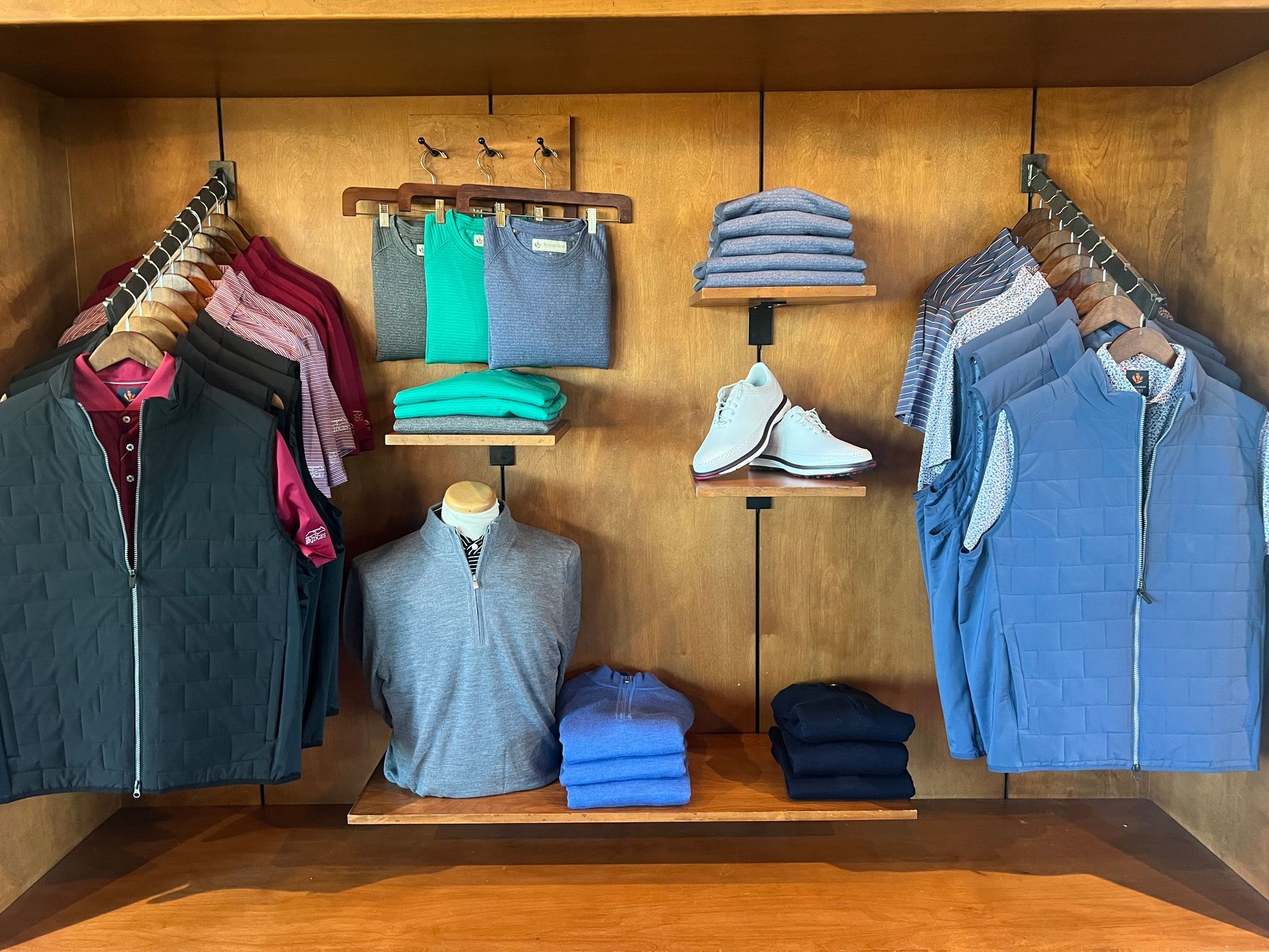 Clothing display in a wooden niche, featuring shirts, pants, vests, and shoes in various colors.