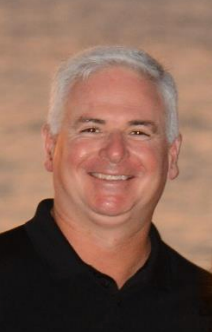 Smiling man with graying hair in a black shirt, outdoors with blurred background.