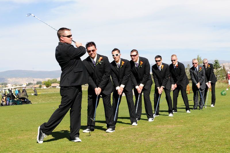 Groomsmen in black suits line up behind a golfer swinging a club on a sunny golf course.