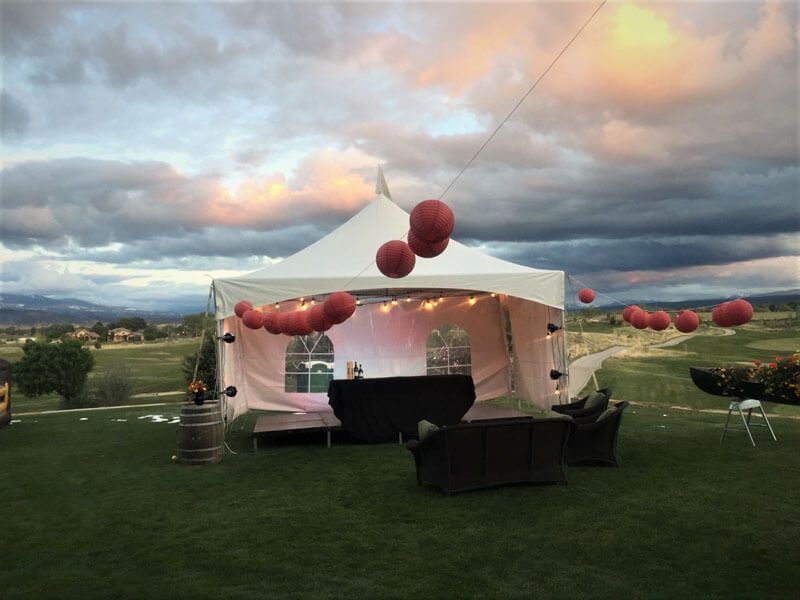 White tent with red balloons, table, and seating on green grass, with a golf course and sunset in the background.