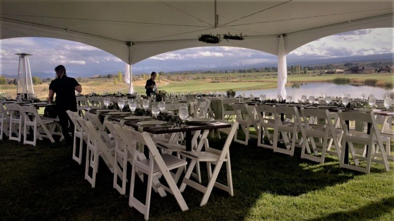 Outdoor event tent with tables set for a meal, overlooking a lake and grassy field; two people preparing.