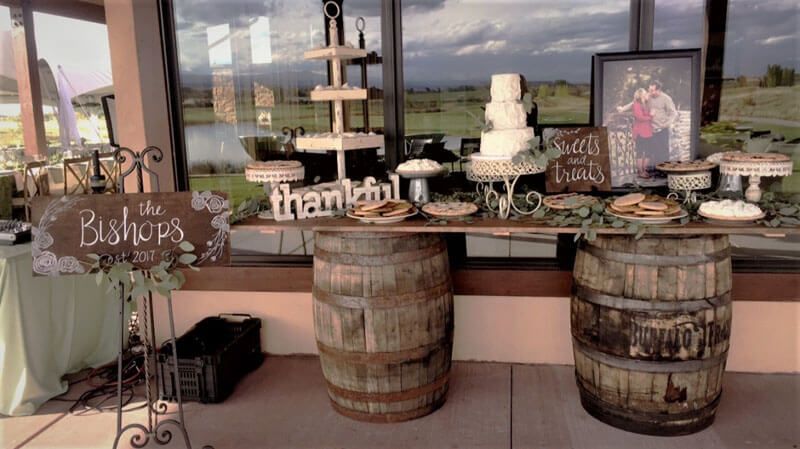 Dessert table with cakes, pies, and floral decor, supported by wooden barrels, with a scenic outdoor backdrop.