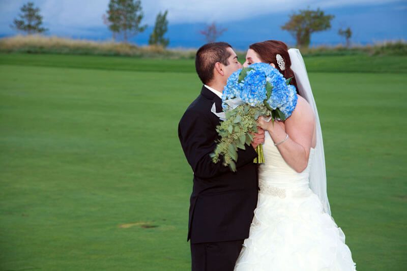 Bride and groom share a kiss, obscured by a blue flower bouquet, on a green golf course.